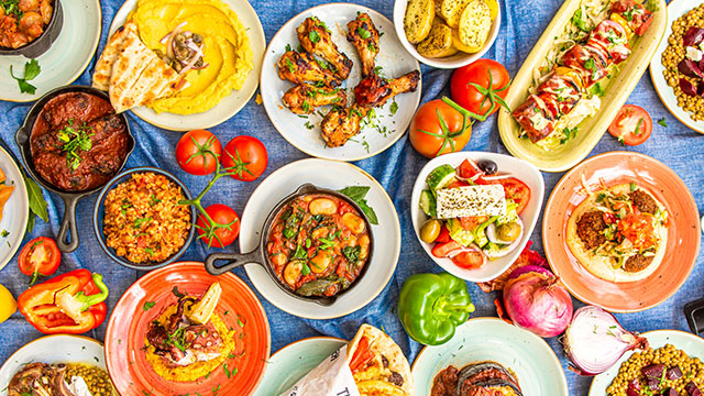 A blue table spread covered with tomatoes and peppers and plates of colourful Greek and Mediterranean food 