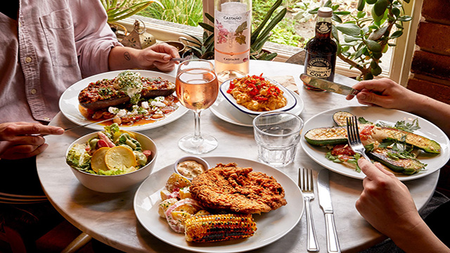Two persons are sitting enjoying a brunch with dishes including Mac and Cheese, plant-based fried chicken amongst others.