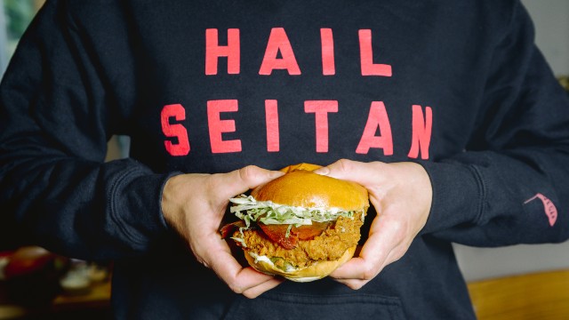 Burger végétalien tenu par une personne vêtue d'un pull noir portant les mots Hail Seitan en rouge, en référence au nom du restaurant, Temple of Seitan.