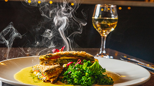 The aubergine schnitzel served with greens is presented on a white plate at The Gate Restaurant with a glass of white wine.