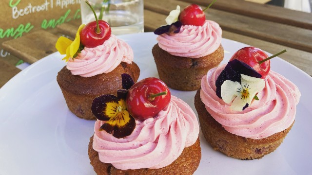 Four cupcakes on a white plate, with pink frosting, yellow flowers and cherries on top.