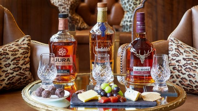 Bottles of whisky on a small table with glasses and plates of cheese, fruit and chocolate. 