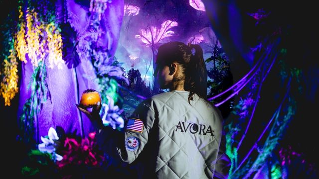 A girl stands in a space suit style jacket holding a cocktail in a magically bright and alien world at the immersive experience Avora in London.