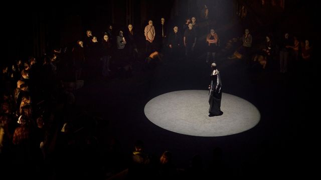 A man in a long black coat stands under a spotlight surrounded by an audience in masks at the burnt city immersive experience in london.