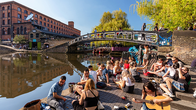 People sit beside Camden Lock on a sunny day, some chatting, some drinking and one playing a guitar - with a bridge and the canal in the background.