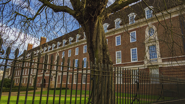 Historic redbrick building with black iron fence running alongside and a large tree without leaves.