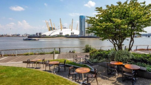 Small wooden circular tables arranged on a curved terrace area by the river Thames with The O2 arena visible in the background.