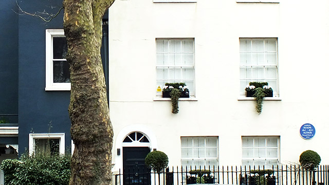 A blue plaque is on a white house with baskets of plants outside the windows.