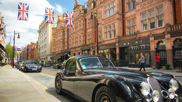 Expensive black cars parked on stylish Mount Street in London's Mayfair, with UK flags hanging between red-brick buildings in the background.