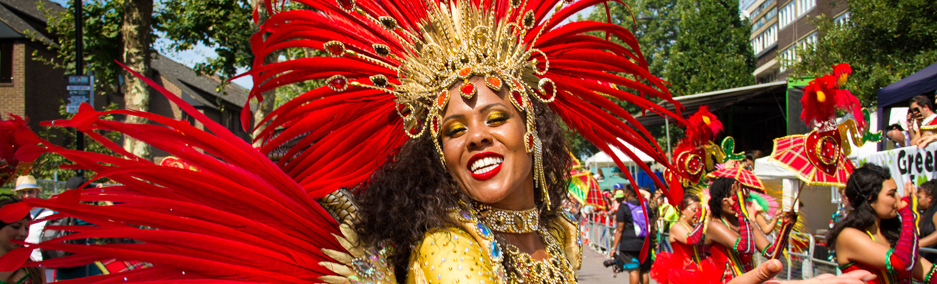 A carnival queen dressed in gold with and red feathers smiling at Notting Hill Carnival on a sunny day