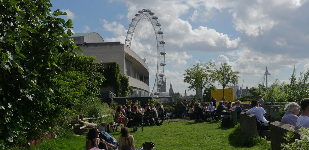 People relaxing on the grass on a bright day at Queen Elizabeth Hall Roof Garden, with the London Eye in the background.