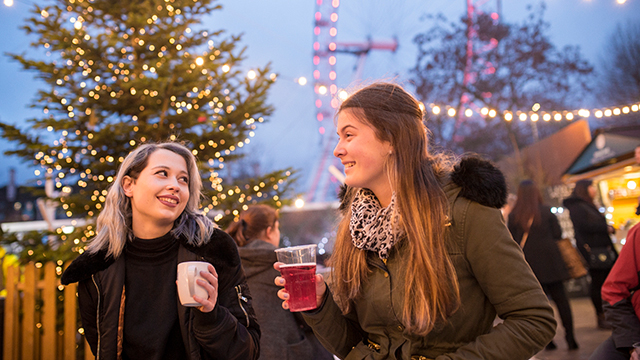 Two women, wearing winter clothes, smile at each other while holding glasses of drink, while fairy lights and the London Eye are illuminated in the background.