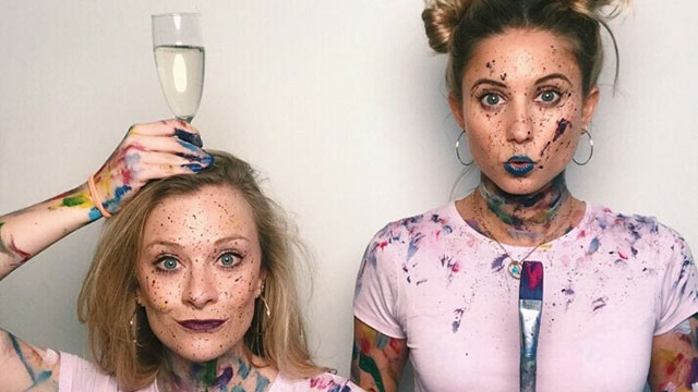 A couple of girls in pink t-shirts covered in paint splashes with one holding a glass of prosescco above her head