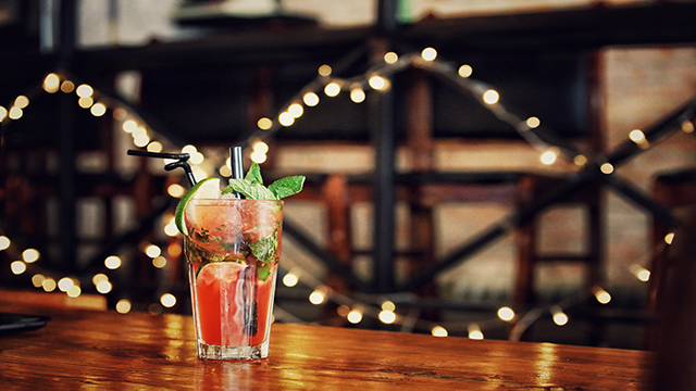 A red cocktail on a brown wooden table with fairy lights in the background.