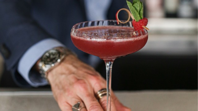 Man in a suit handing over a red cocktail decorated with a raspberry on a cocktail stick.
