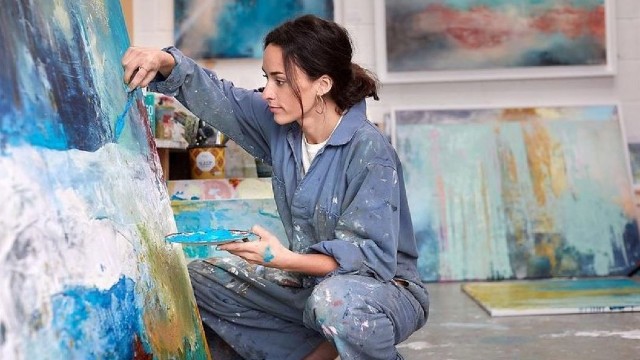 Einen Frau malt mit blau, weiß und gelb auf eine Leinwand und Gemälde stehen im Hintergrund.