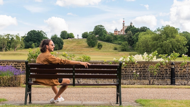 Man sitting on a bench in Greenwich Park with the Royal Observatory in the distance on a sunny day.