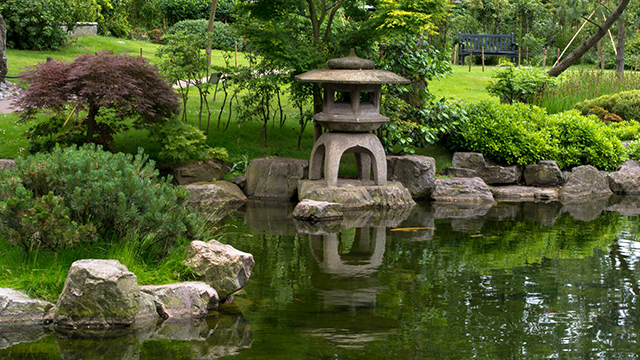 A Japanese-style sculpture reflects in the water of a pond in Kyoto Garden, with acers surrounding the pond.