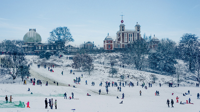 The hill at Greenwich Park covered in snow, with many people standing around, and the Royal Observatory in the background.