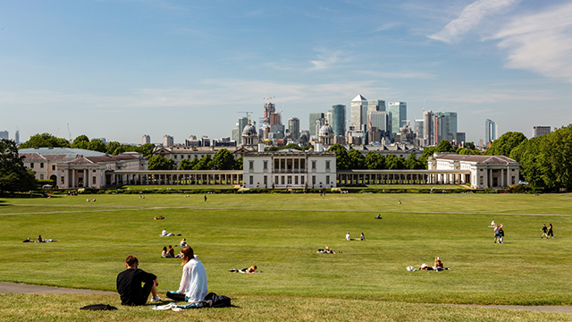 Groups of people sit on the hill of Greenwich Park overlooking the Old Royal Naval College and the London skyline. 