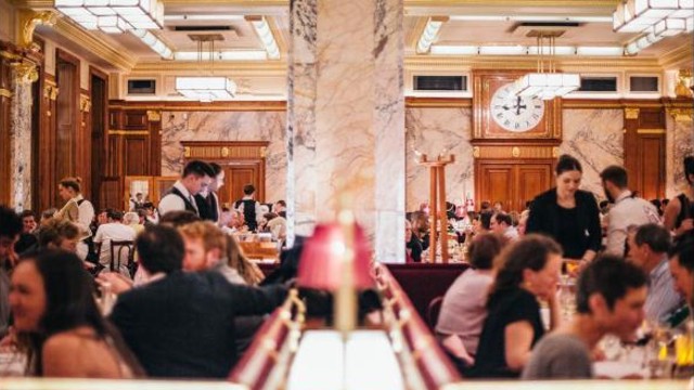 Numerous elegant waiters serve tables in a busy Art Deco restaurant full of clients.
