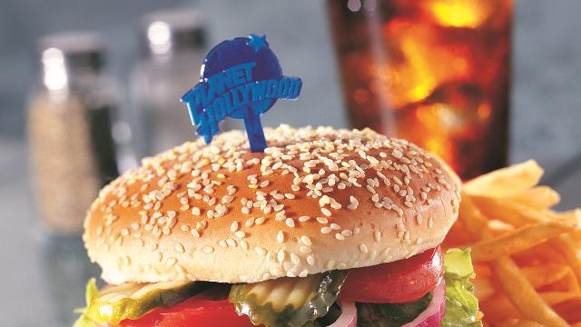 A tasty American classic burger with pickles and tomato is held together by a Planet Hollywood blue toothpick, served with fries and a fizzy drink in the background .