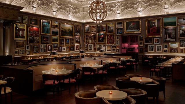 A grand dining room with beautiful wooden floors is lit with soft candle lights; its walls are decorated with an incredible collection of paintings.
