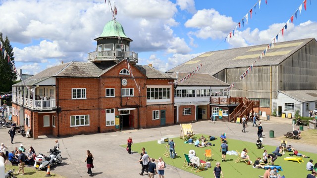 People are sitting on the grass in front of Brooklands Museum on a sunny day.