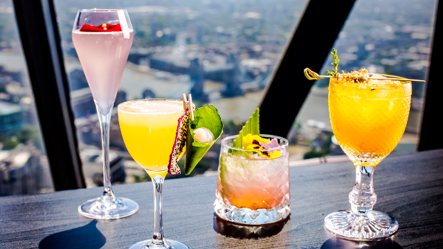 Four colourful cocktails in various glasses, decorated with fresh fruit and edible flowers, sat against a backdrop of the view from The Gherkin.