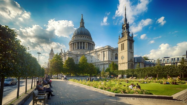 A view of St Paul's Cathedral taken from the south east side. There are people sitting on benches and on the grass outside the cathedral in the foreground.