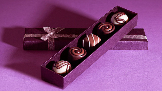 A purple box  with a ribbon containing five different chocolates on a purple table
