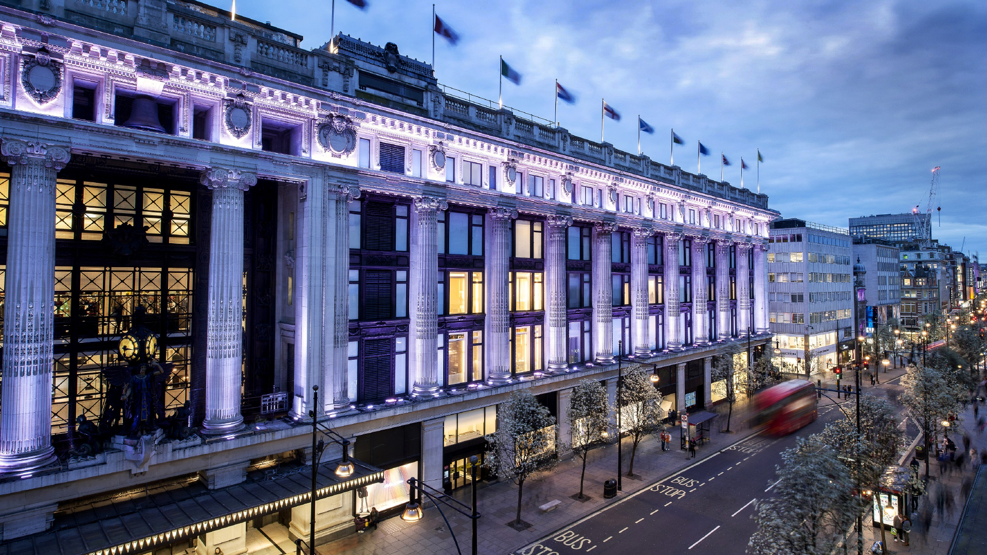 A panoramic view from high of the facade of Selfridges, lit up in purpley-white lights, with Oxford Street below.