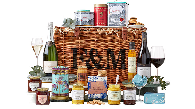 A wicker basket hamper with the logo "F&M" writte on it in black, with various jars of jams, pickles and condiments, plus bottles of wine and sparkling wine, tins of biscuits and other food on and around the hamper.