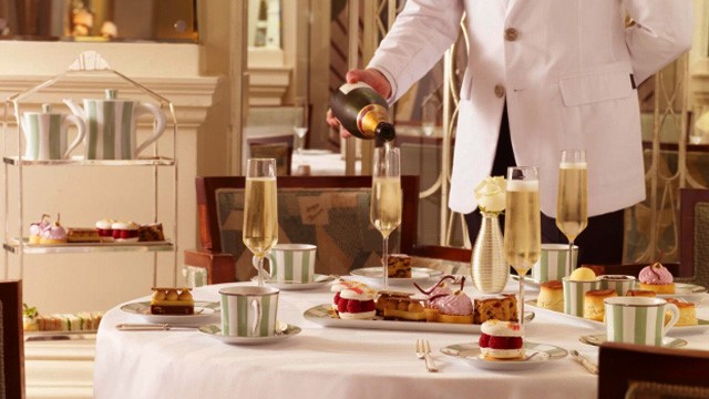 Ein Kellner gießt beim Nachmittagstee Champagner in ein Glas, während Kuchen und Tassen auf dem Tisch stehen.