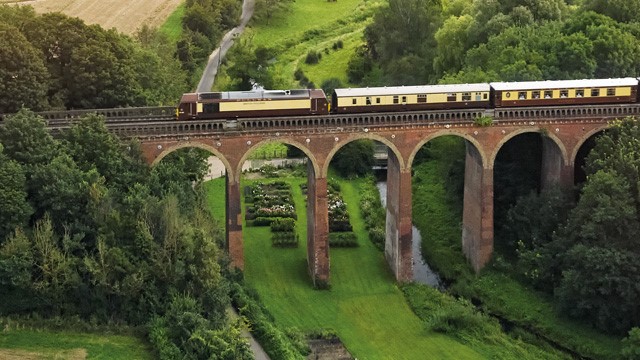 Un luxueux train Pullman British Belmond circulant sur un pont viaduc entouré d'une campagne verdoyante.