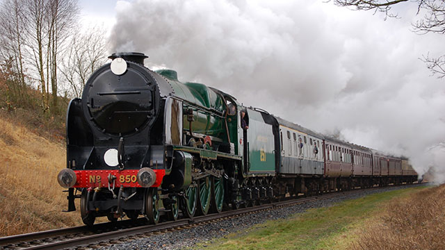 A black and green steam train billows out steam as it moves on tracks towards the camera, with grass verges to the side.