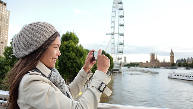 A lady, wearing a beanie hat and a coat, takes a photograph with her phone, with the London Eye in the background.