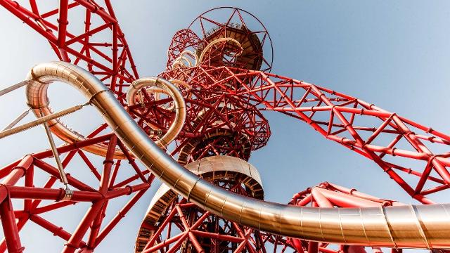 Looking up from the ground at the twisting red structure of the ArcelorMittal Orbit, with the winding grey tunnel slide intertwined, on a sunny day.