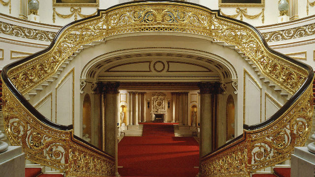 Vue du grand escalier couleur or du palais de Buckingham et une moquette rouge recouvre le sol. 