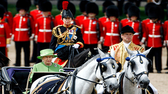 The queen wearing a green hat and dress sits on a carriage during trooping the colour.