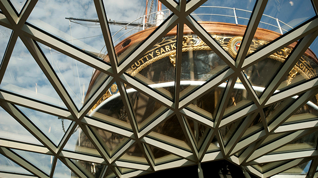 Looking up from beneath the hull of Cutty Sark - with the crisscross pattern of the glass ceiling above and the stern of the black and gold ship beyond