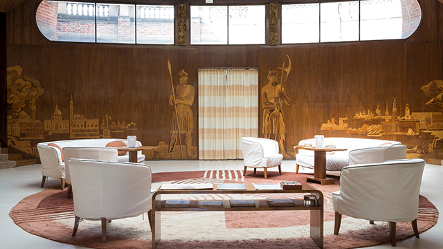 The entrance hall of Eltham Palace, with art-deco wood panelling featuring gold-coloured designs of soldiers and landscapes, white chars and a circular pink rug.