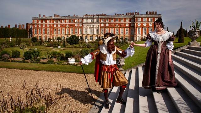Deux personnages en costumes d'époque se tienne au premier plan et semble se faire la court, avec une vue sur le palais de Hampton Court à l'arrière plan.
