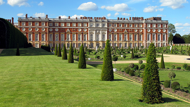 A row of trees and a neatly kept lawn in the foreground leads towards the red brick facade of Hampton Court Palace in the background.