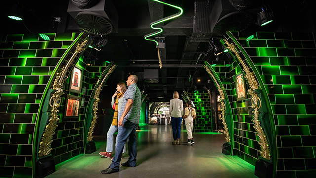 Zwei Personen betrachten in der Harry-Potter-Fotoausstellung gerahmte Bilder an einer gekachelten Wand, die in leuchtend grünes Licht getaucht ist, mit einem neongrünen, sich windenden Licht darüber. 