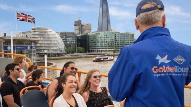 A group of tourists sit on an open top bus going past The Shard and across the river Thames with a local guide.