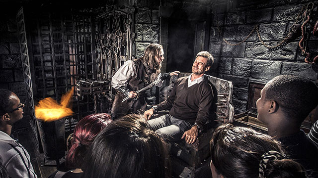 Ein verängstigter Mann sitzt in einem Stuhl während ein andere Mann in einem mittelalterlichen Kostüm in foltert