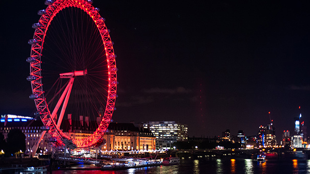 The London Eye, illuminated in red, at night, with the river Thames in the foreground.