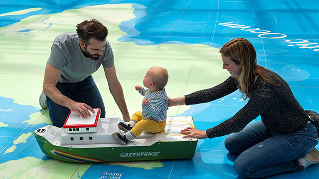 A mother and father guiding their baby sitting on a little boat over a green and blue map of the world