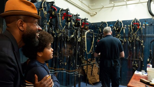 A father and son watch an expert of the royal mews as he tends to horse bridles.